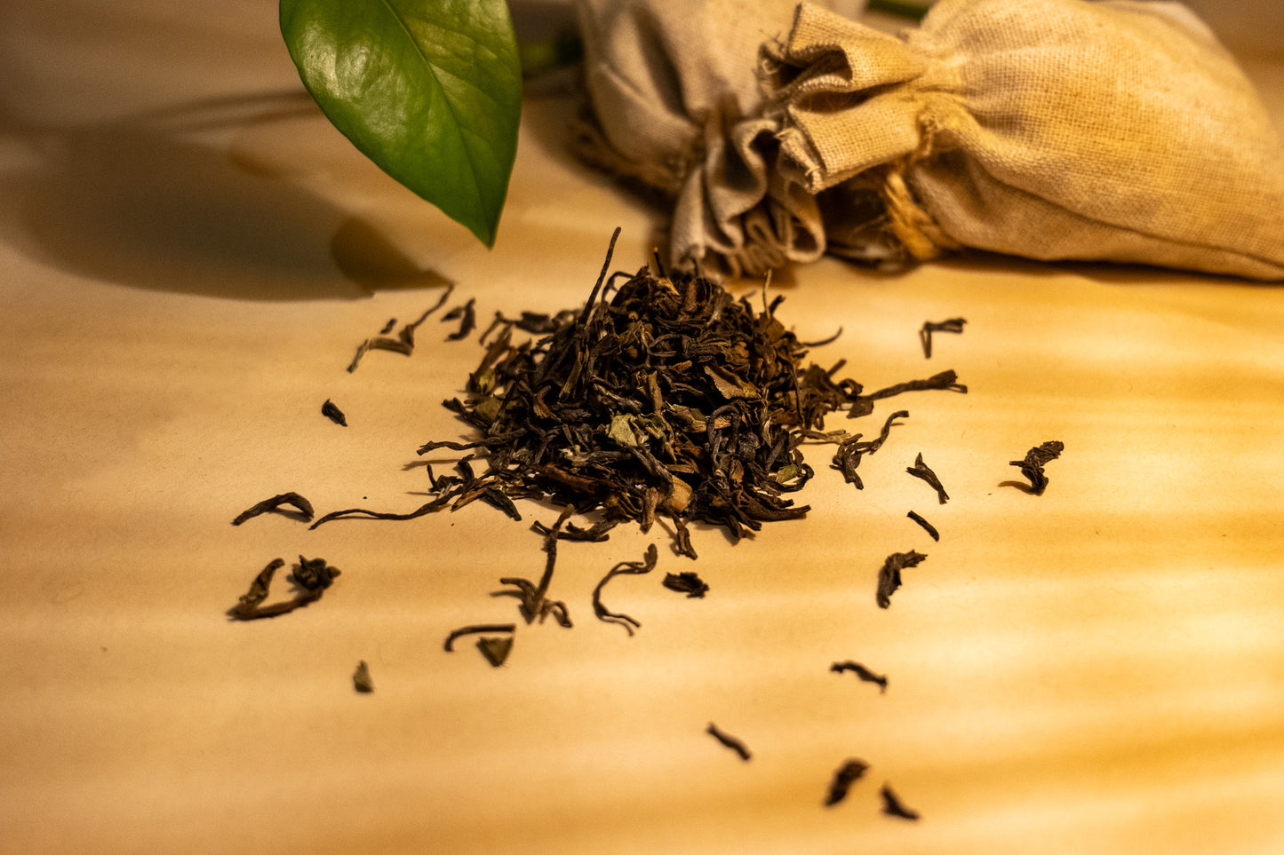 A pile of dry, rolled Darjeeling black tea leaves on a light surface, with a few leaves scattered around. There's a green leaf and a burlap sack in the background, suggesting a natural and organic setting.