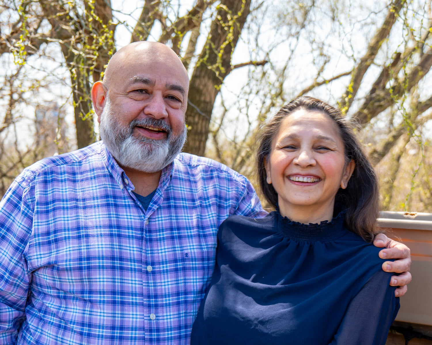 A smiling Indian couple on a sunny day. The man is bald with a white-grey beard, and is wearing a blue and purple plaid shirt with the top button undone to show a blue V-neck T-shirt underneath. The woman is wearing a navy blue blouse with sheer sleeves and a ruffled collar.