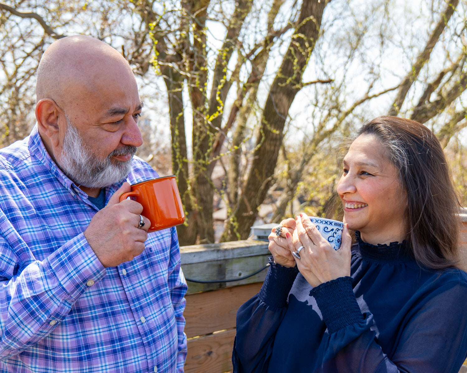 An Indian couple holds their mugs and smiles at each other on a sunny day. The man is bald with a white-grey beard, and is wearing a blue and purple plaid shirt with the top button undone to show a blue V-neck T-shirt underneath. His mug is a rust-orange color. The woman is wearing a navy blue blouse with sheer sleeves and a ruffled collar. Her mug is blue and white with floral patterns.