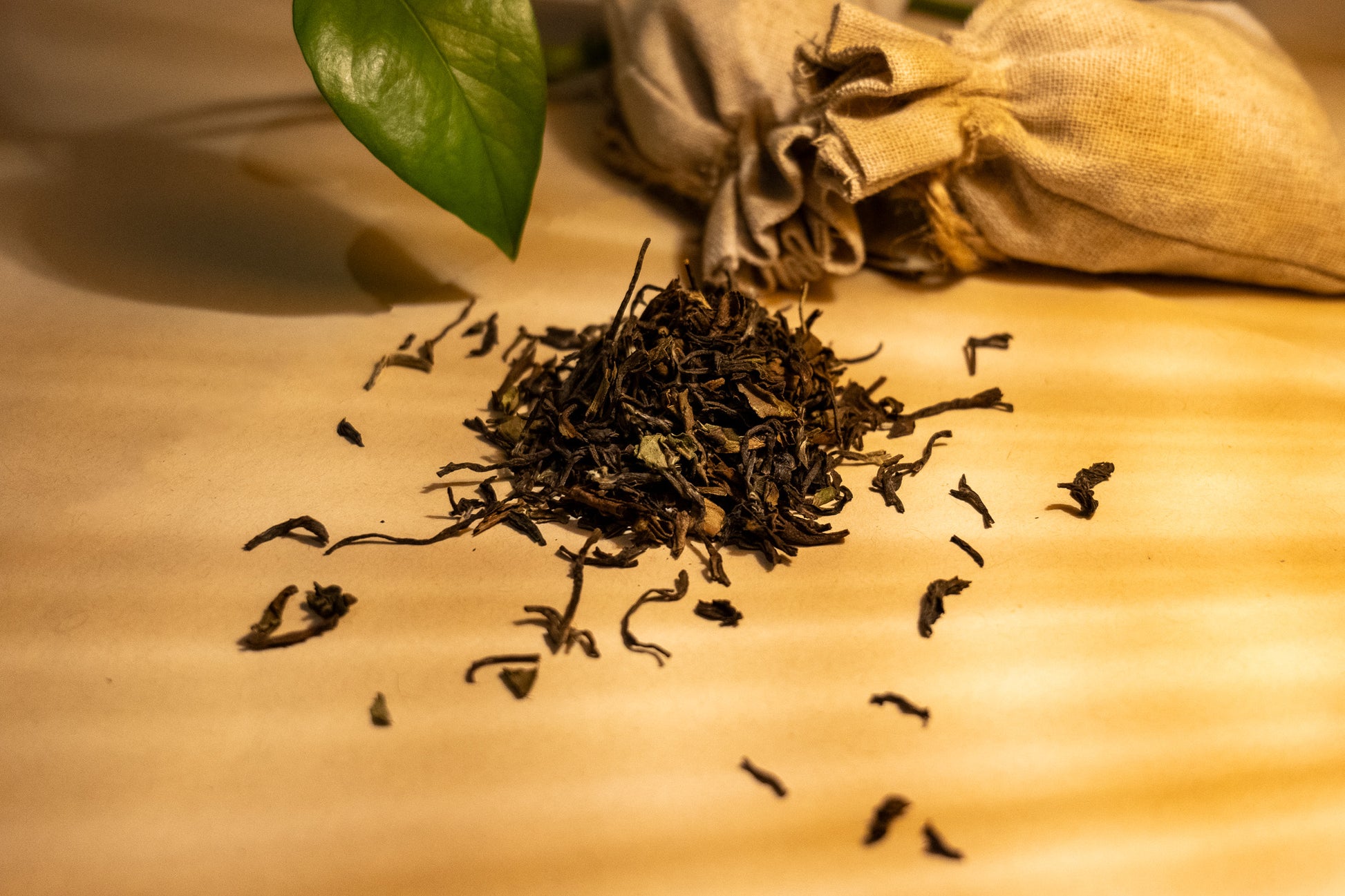 A pile of dry, rolled Darjeeling black tea leaves on a light surface, with a few leaves scattered around. There's a green leaf and a burlap sack in the background, suggesting a natural and organic setting.