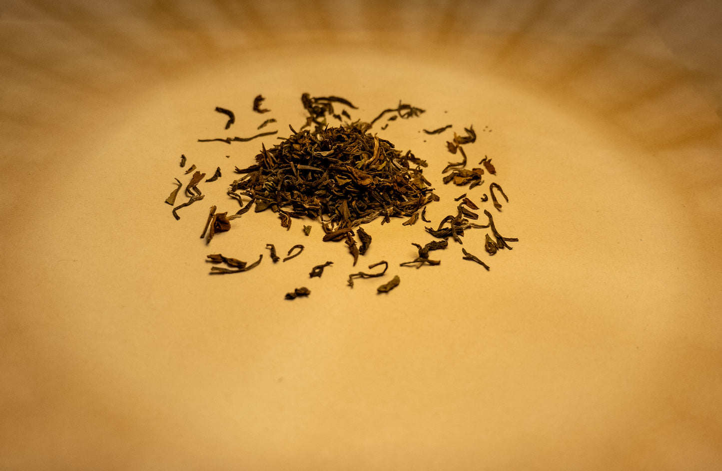 A pile of dry, rolled Darjeeling black tea leaves on a light surface, with a few leaves scattered around. The background is an orange-brown color, suggesting a natural and organic setting.