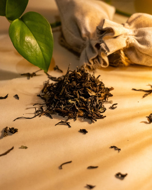 A pile of dry, rolled Darjeeling black tea leaves on a light surface, with a few leaves scattered around. There's a green leaf and a burlap sack in the background, suggesting a natural and organic setting.