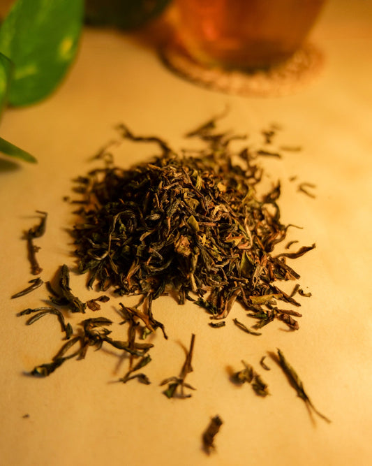 A pile of dried, whole leaf Darjeeling black tea on a light background, with a teacup and green leaves in the background.