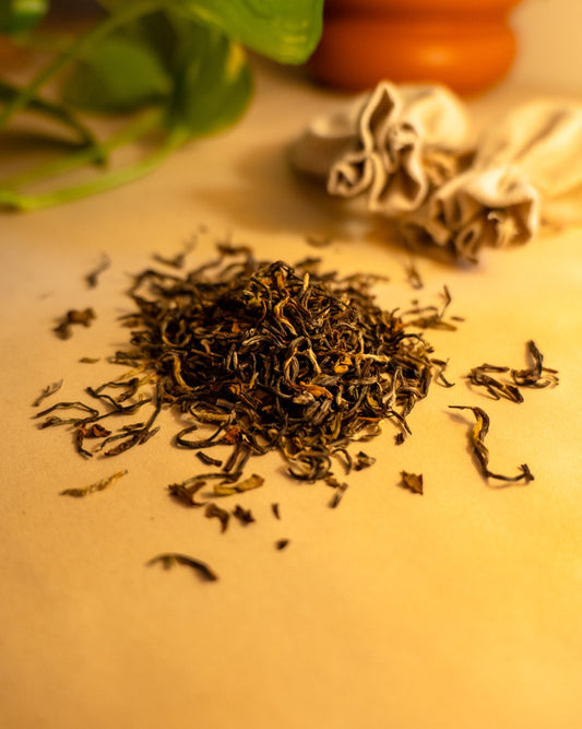 A pile of dried, rolled black tea leaves on a wooden surface, with a blurred background suggesting a cozy indoor setting. Behind the pile are tea pouches, leaves, and a clay pot
