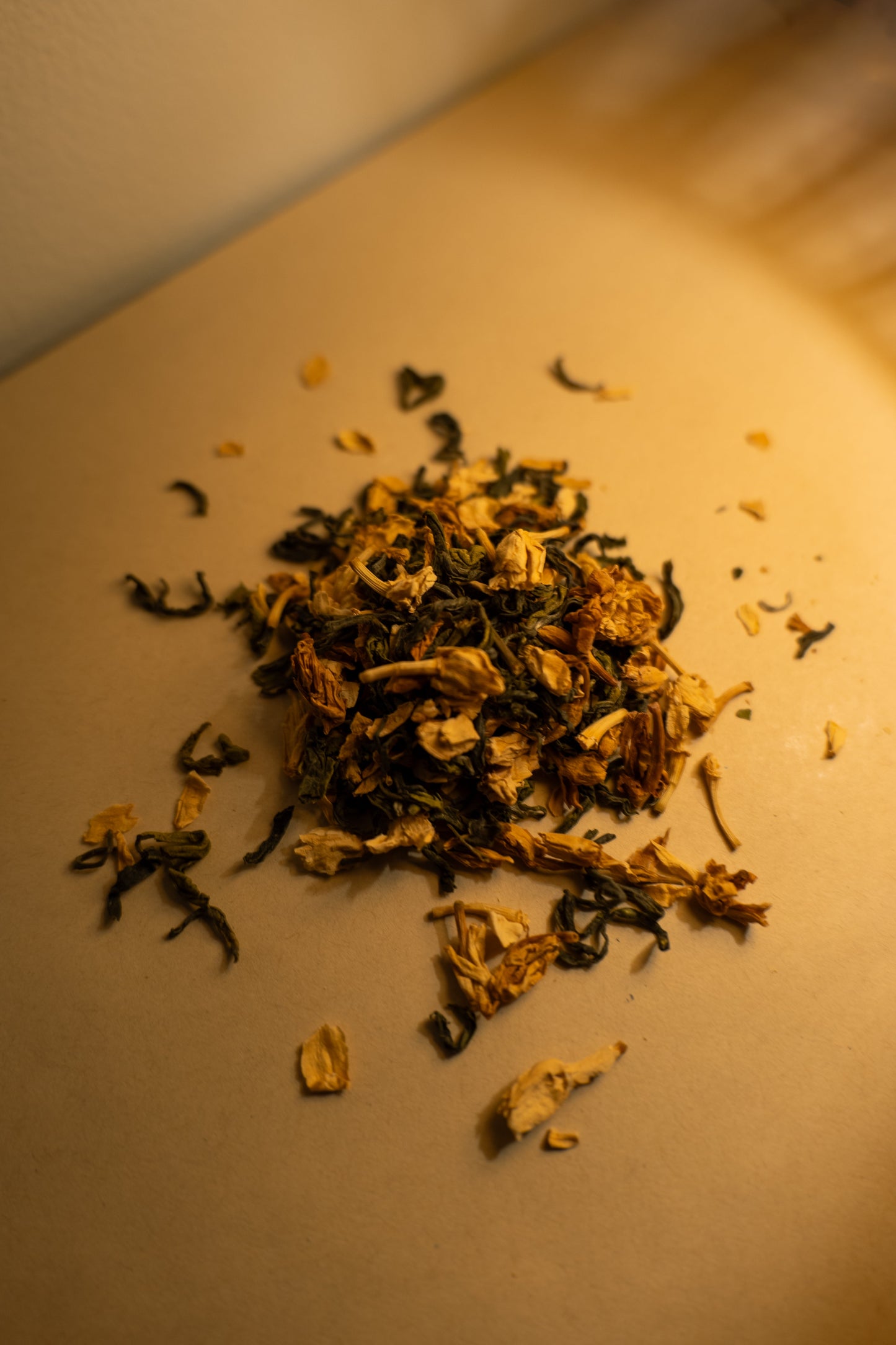 A scattered pile of dried jasmine green tea leaves and jasmine flowers on a light surface with a white background. The photo is shot at a tilted angle and light is spilling onto the pile from somewhere behind and to the side off-camera.