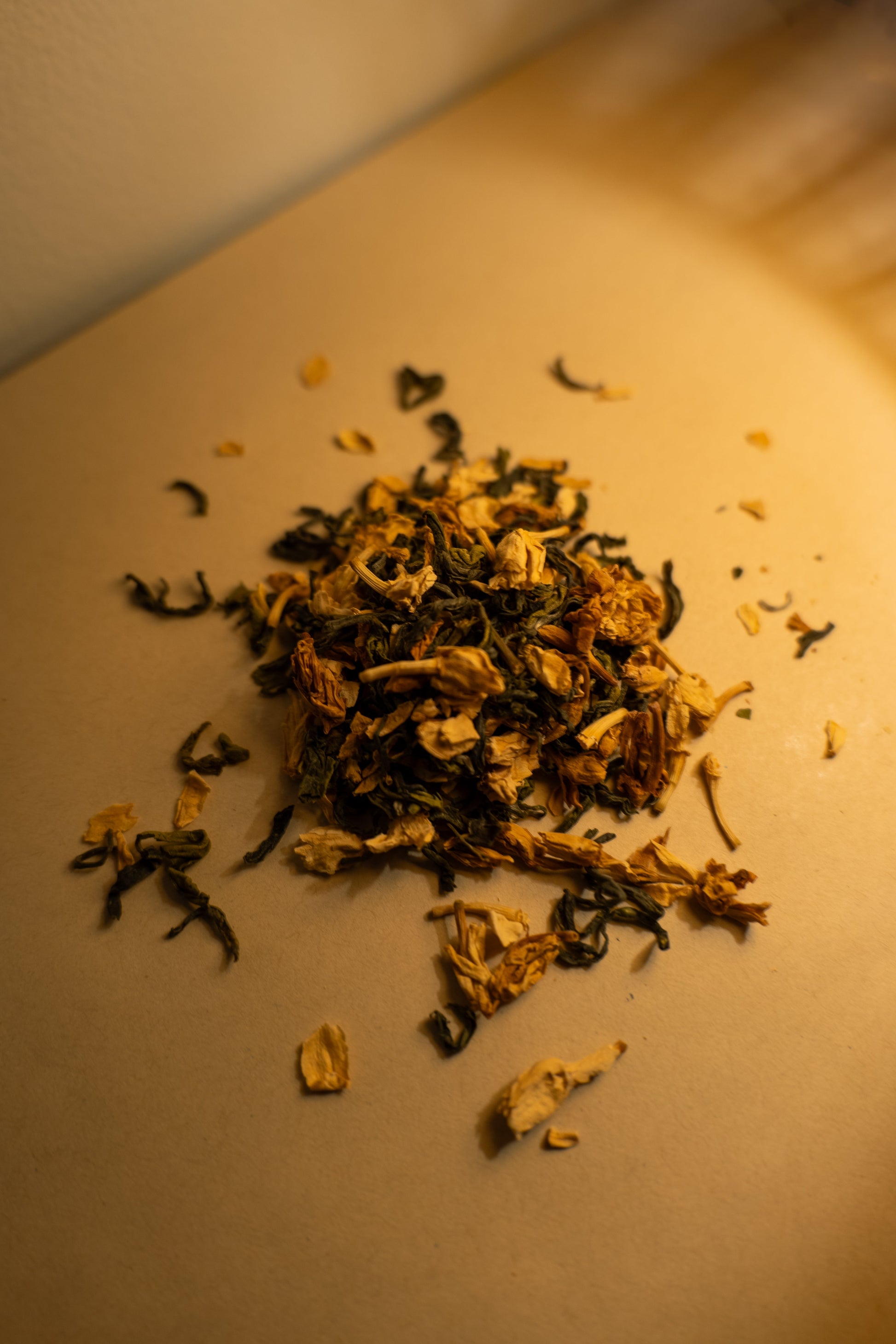 A scattered pile of dried jasmine green tea leaves and jasmine flowers on a light surface with a white background. The photo is shot at a tilted angle and light is spilling onto the pile from somewhere behind and to the side off-camera.
