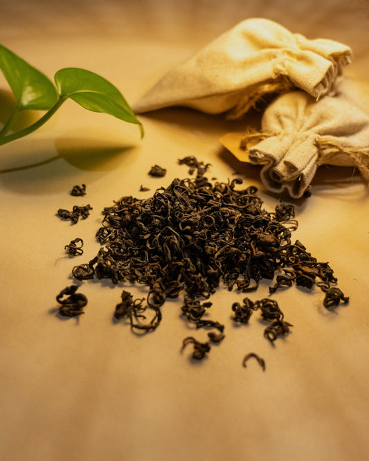 A pile of withered black tea leaves on a wooden surface, with a green leaf and a cloth bag in the background.