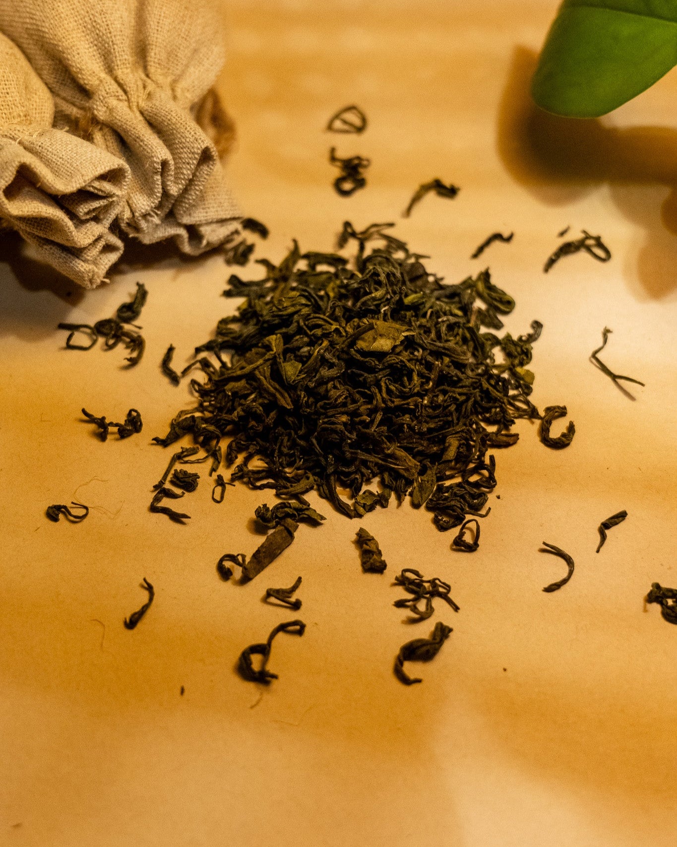 A scattered pile of dried green tea leaves on a brown surface, with a tea bag and a green leaf in the background.