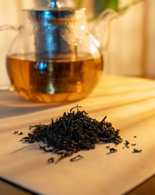 A pile of loose tea leaves in front of a clear teapot filled with tea, on a wooden surface with a white background. The lighting and coloring is warm, suggesting a natural and organic setting.