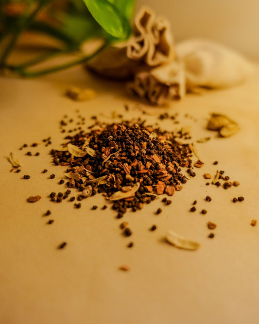 A variety of spices including cinnamon, cardamom, cloves, ginger, and black pepper scattered on a surface, with a tea bag and leaves in the background.