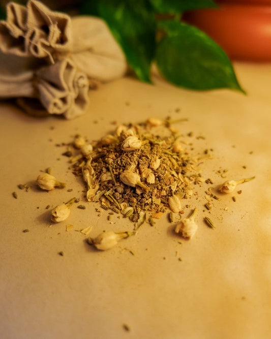 A variety of dried herbs and spices, including saffron, cinnamon, cardamon, clove, black pepper, ginger, fennel, jasmine buds, are scattered on a surface, with a pouch and leaves in the background.
